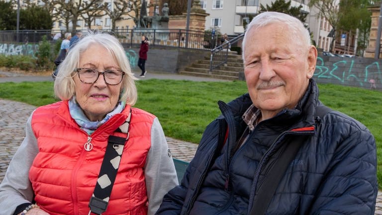 Eine ältere Dame mit Brille und pinker Jacke und ein älterer Herr mit dunkler Jacke sitzen auf einer Bank und schauen in die Kamera.  
