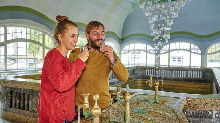 Ein Paar trinkt Solewasser aus den Hähnen in der Brunnenhalle in Bad Münster am Stein